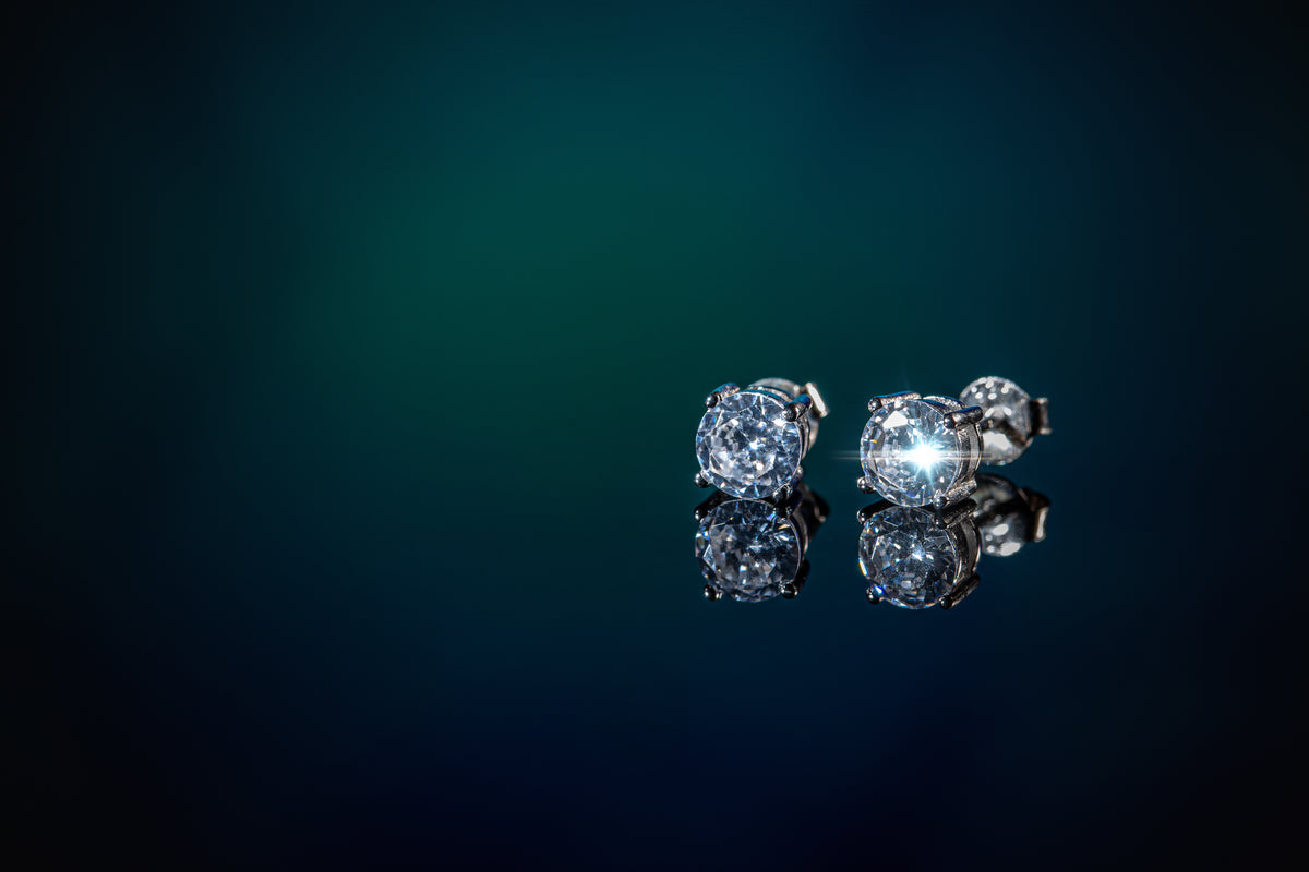 A pair of round-cut diamond stud earrings in a silver-toned setting, displayed on a reflective dark blue surface against a dark teal background.
